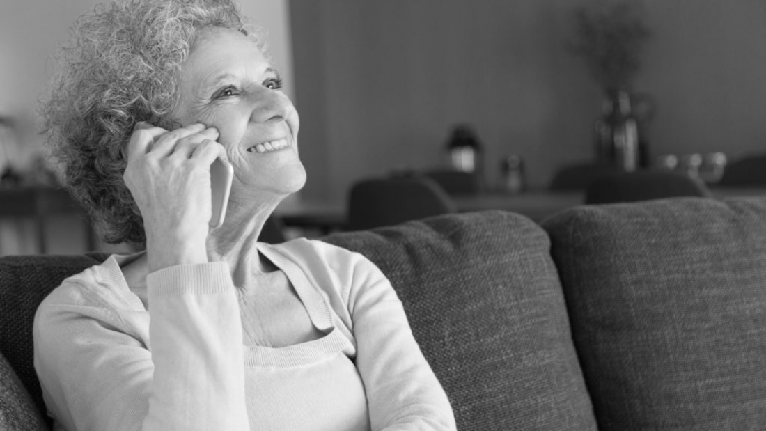 Close-up of happy senior woman talking on smartphone. Smiling grandmother sitting on couch at home and answering call. Elderly people and communication concept